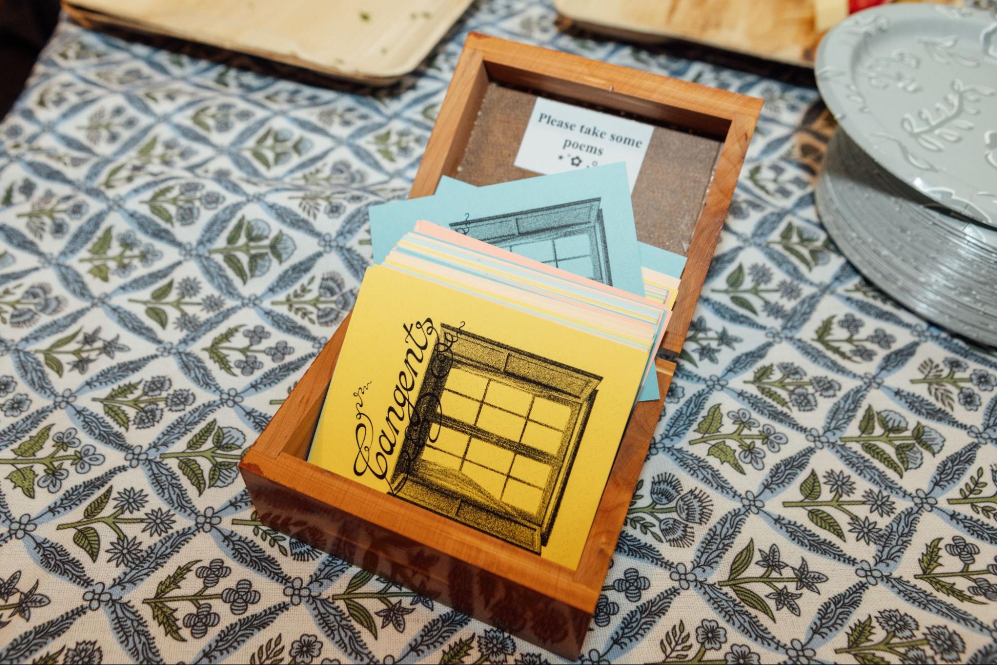 A wooden box contains colorful cards stacked inside, with a note saying "Please take some poems." The cards feature designs of windows and decorative letters. The box sits on a patterned tablecloth with floral motifs, and some silver plates are visible in the background.
