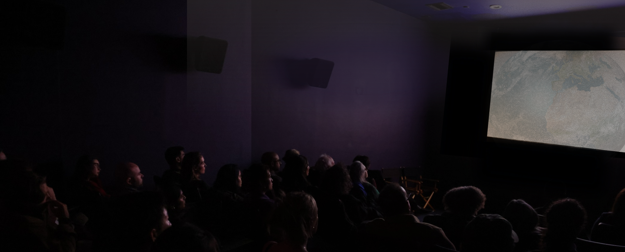 A dimly lit movie theater filled with an audience watching a film. The screen shows a textured image, possibly a landscape or abstract design. People are seated in rows, some appearing focused on the screen, while others are relaxed or deep in thought. The walls are dark, enhancing the cinematic atmosphere.