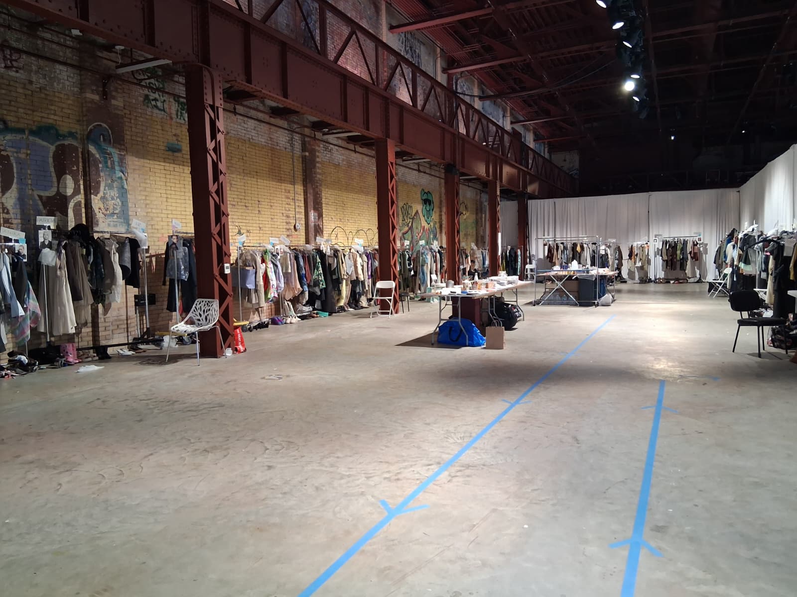 An expansive, industrial space featuring a concrete floor and exposed brick walls. On one side, rows of clothing are displayed on hangers and racks. There's a table with scattered items in the center, and a white curtain at the far end. Blue tape lines the floor, creating pathways through the area.