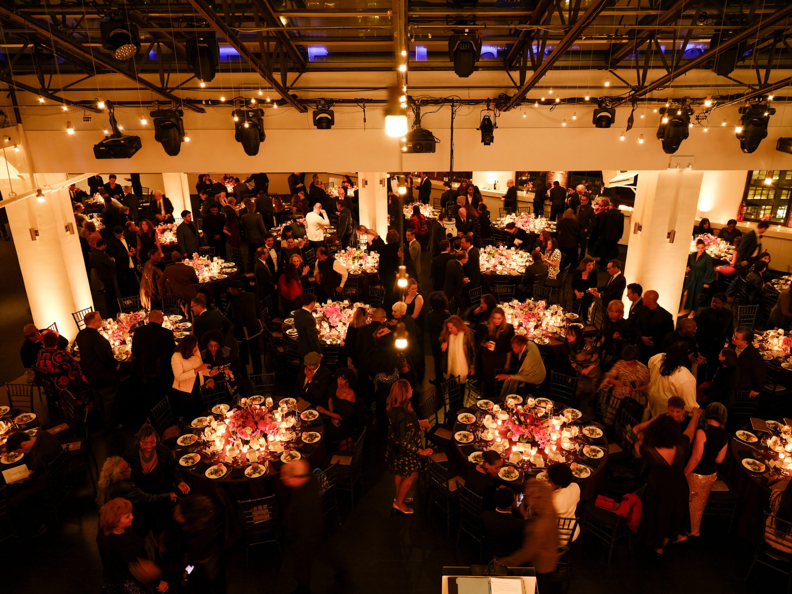 A spacious banquet hall filled with elegantly dressed guests socializing and dining. Tables adorned with floral centerpieces and candles display plates of food. The area is illuminated by warm lighting and decorative string lights, creating a festive atmosphere. Guests are seen mingling, with some seated and others standing, contributing to a lively gathering.