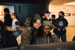 A group of people are gathered in a tech space, engaging with computers and wearing headphones. A woman with long black hair and glasses leans over a man with glasses and headphones, who is focused on the screen. In the background, others are interacting, with some facing away from the camera. The environment appears modern and collaborative, with soft lighting and a digital display on the wall.