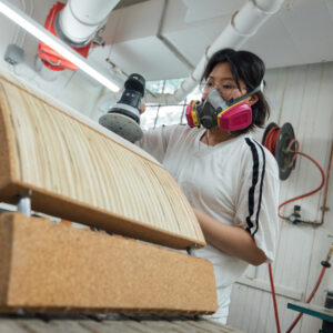 A person wearing a mask and safety glasses is sanding a piece of wood with a power sander. They are in a workshop with various tools and machinery visible in the background. The wood has visible grooves and layers, indicating it's part of a larger project.