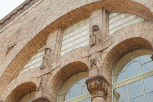 Close-up view of a brick facade featuring two stone sculptures of figures adorned in long robes, each standing beside an arch-shaped window. The figures are positioned on either side of the central arch, holding a banner. Decorative stonework details can be seen beneath the figures, highlighting intricate designs. The overall architectural style suggests a historical significance.