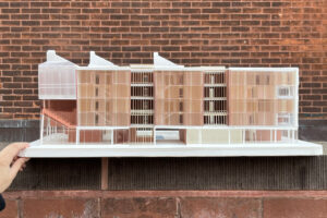 A hand holds a scale architectural model of a building, showcasing various transparent and colored materials. The model features distinct sections with a contemporary design, including layered horizontal elements and a sloped roof. The background consists of a textured red brick wall, highlighting the model's details.