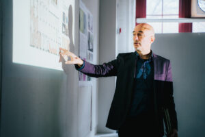 A man in a suit gestures toward a projected presentation on a wall. He has a serious expression, and the projection displays architectural diagrams and images. The room has large windows with light streaming in, creating contrasting shadows on the wall. Behind him are sheets of paper pinned to the wall.