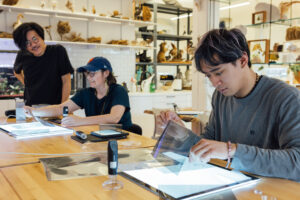Three individuals are engaged in a hands-on activity at a wooden table in a well-lit workspace. One person is using a lightbox to examine a transparent sheet; another person, wearing a cap and glasses, is pointing at materials, while the third person stands in the background, observing. Various tools and supplies are scattered on the table, and shelves in the background hold natural specimens and other items.