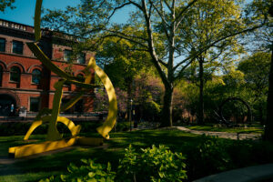 A vibrant park scene featuring a large yellow abstract sculpture in the foreground, surrounded by lush green grass and foliage. Tall trees with bright green leaves provide shade, while a brick building is visible in the background. Additional greenery includes flowering trees, and a black circular sculpture is positioned to the right. The sky is clear blue, indicating a sunny day.