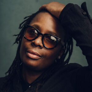 A person with dark, shoulder-length dreadlocks is posing, looking directly at the camera. They are wearing large, stylish sunglasses and have one hand resting on their head. The background is a softly blurred green tone, emphasizing the subject's facial features and expression.