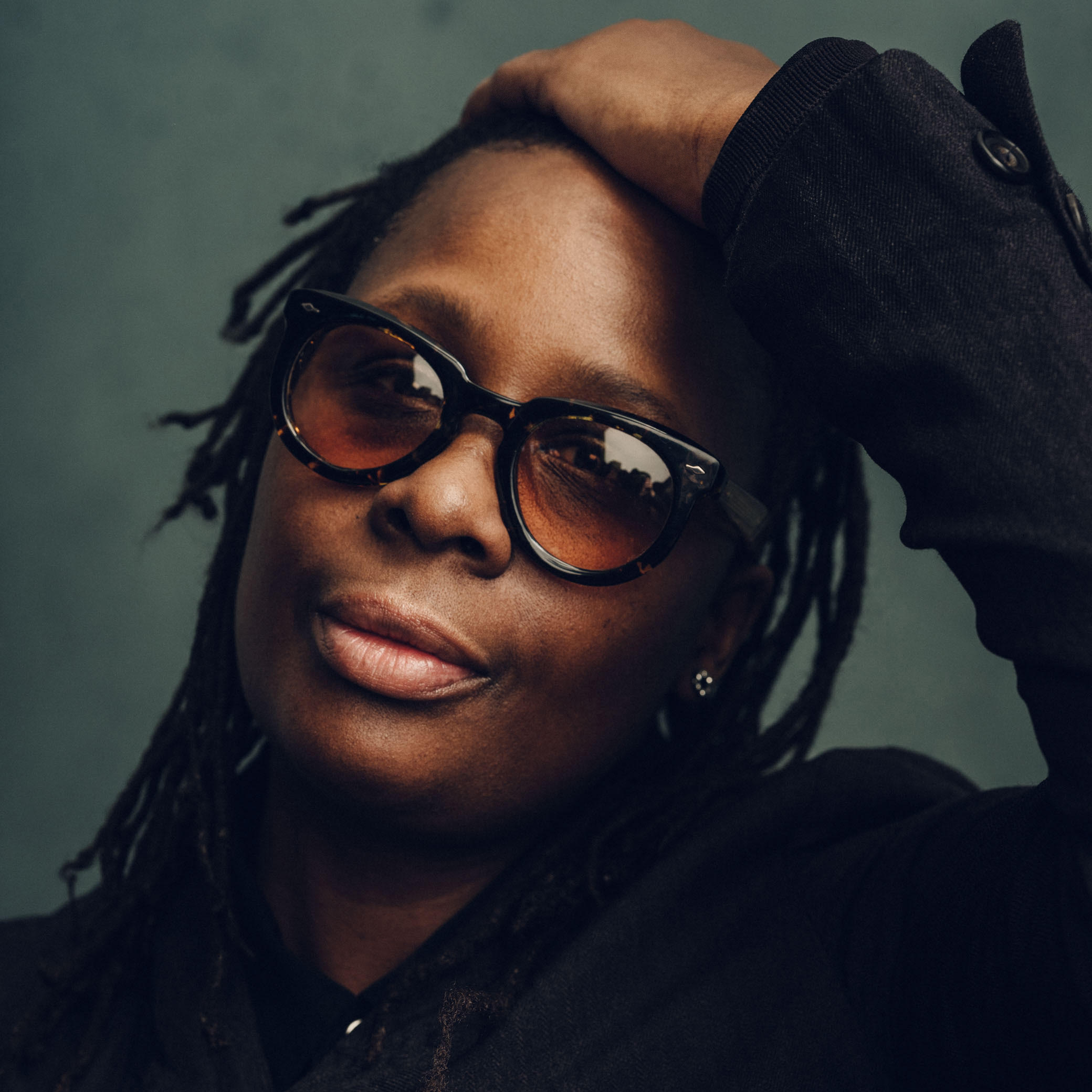 A person with dark, shoulder-length dreadlocks is posing, looking directly at the camera. They are wearing large, stylish sunglasses and have one hand resting on their head. The background is a softly blurred green tone, emphasizing the subject's facial features and expression.