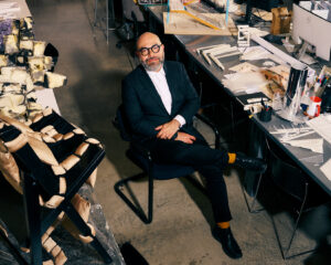 A seated man with glasses and a beard is positioned in a modern studio. He wears a black suit and a white shirt, with orange socks contrasting against his black shoes. Surrounding him are various design materials and models, including a sculptural piece and multiple architectural sketches spread across the table, indicating a creative workspace.