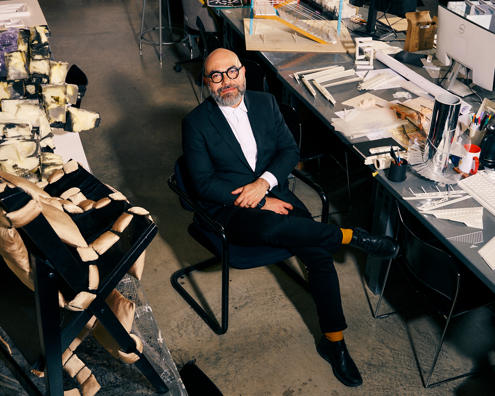 A seated man with glasses and a beard is positioned in a modern studio. He wears a black suit and a white shirt, with orange socks contrasting against his black shoes. Surrounding him are various design materials and models, including a sculptural piece and multiple architectural sketches spread across the table, indicating a creative workspace.