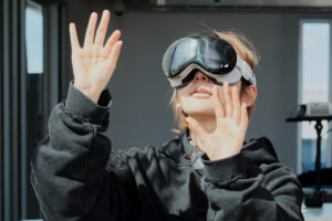 A person wearing futuristic virtual reality goggles raises both hands, with one hand open and fingers splayed. They are dressed in a black hoodie and appear to be engaged with a virtual experience. The background is bright and minimalistic, suggesting a modern environment.