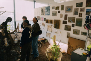 Three individuals are standing in an art studio filled with artwork and sculptures. A prominent sculpture resembling a tree figure is in the foreground, while the walls display various drawings and mixed media pieces, depicting nature and landscapes. The room has natural light coming through large windows, and plants are integrated into the space, contributing to an earthy atmosphere.