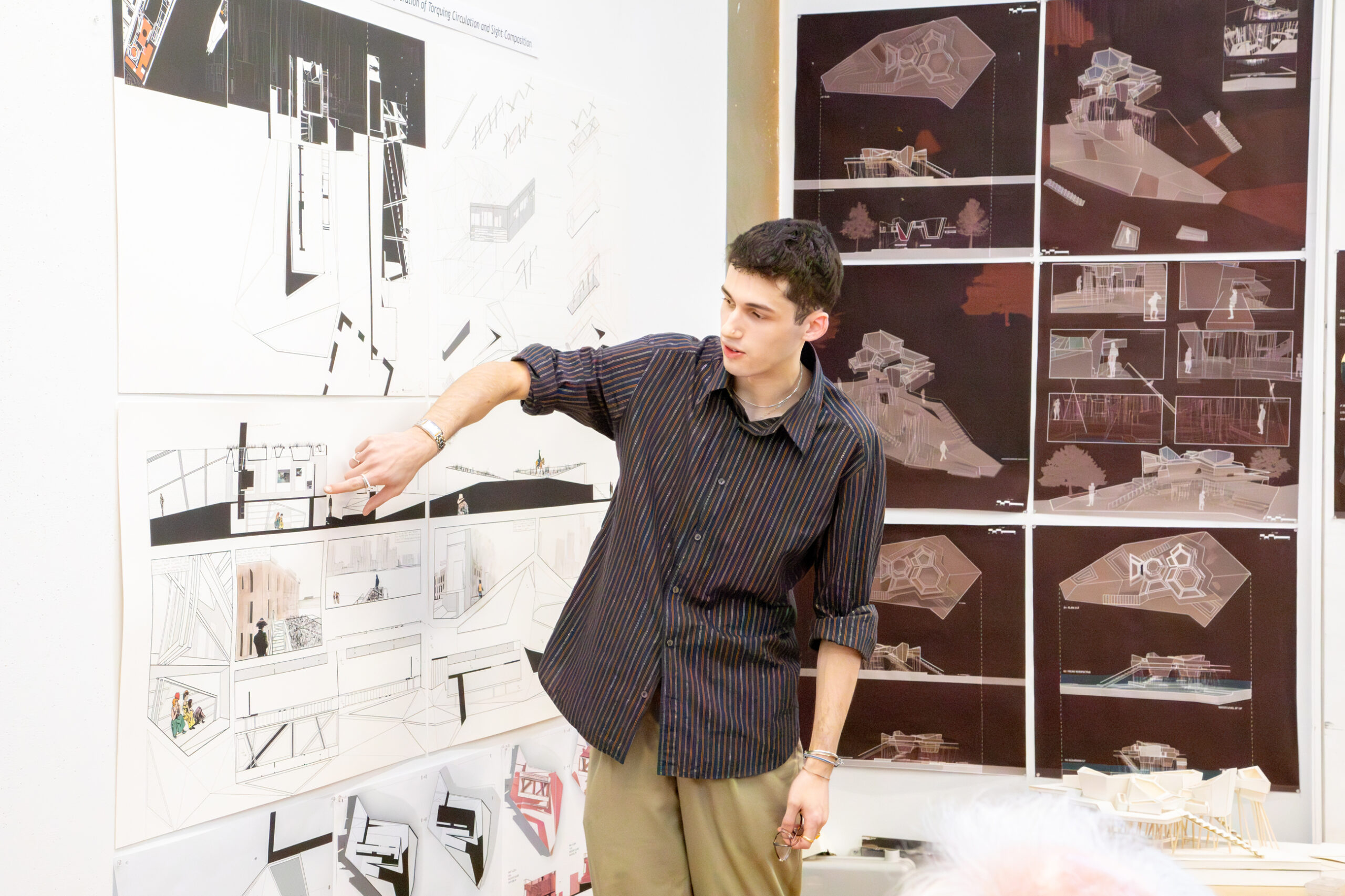 A young man with dark hair, wearing a striped black and blue shirt, stands in front of several architectural drawings pinned to a wall. He gestures towards one drawing with his right hand, explaining it to an audience. The drawings depict various architectural designs, floor plans, and model representations. In the foreground, out of focus, there's a glimpse of a person's head with white hair.
