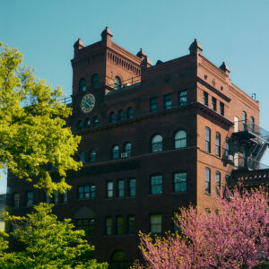 A historic red brick building with ornate architectural details, featuring a clock on its facade. The building has multiple large windows and a fire escape on the right side. In the foreground, vibrant green trees and blooming pink flowers add color to the scene under a clear blue sky.