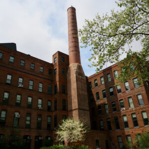 A tall, red brick smokestack rises from the center of an old industrial building complex. The building features multiple large windows with some air conditioning units visible. Surrounding the structure are green trees and plants, indicating a landscaped courtyard. The sky above is partly cloudy.