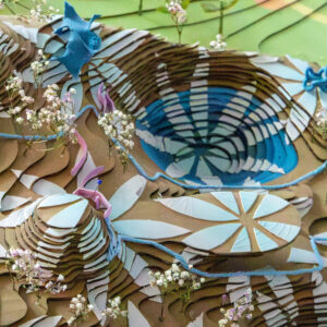 An abstract top-down view of a colorful, layered model depicting a landscape. The model features swirling textures and patterns resembling hills, water features, and flowers. Light blue, dark blue, and pink elements create depth and contrast against the brown base. Small white flowers are scattered throughout, adding a natural touch to the artistic design.
