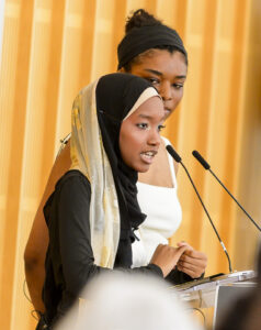 A young woman wearing a black hijab and a long-sleeved shirt speaks at a podium, with a microphone in front of her. Behind her, another woman in a white dress appears to be listening. The background features wooden paneling, creating a formal setting for the speech.