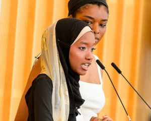 A young woman wearing a black hijab with a patterned scarf speaks into a microphone, appearing passionate. Behind her, another woman in a white sleeveless top looks on attentively. The background features wooden paneling, suggesting an indoor setting, possibly a conference or event.