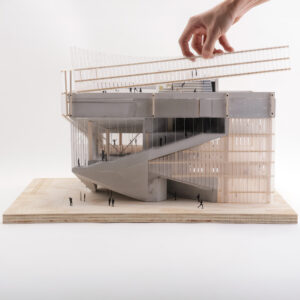 A hand is adjusting a clear architectural model of a modern building, which features a prominent curved staircase, large glass windows, and layered horizontal elements at the top. Small human figures are placed around the model, giving a sense of scale. The base of the model is made of wood and the overall design appears contemporary and complex.