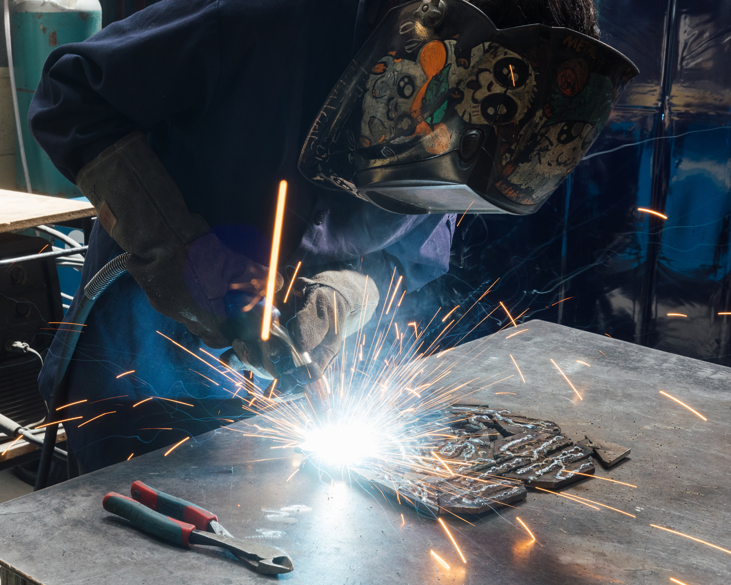 A welder wearing a protective helmet and gloves is focused on a metal piece, producing bright sparks as they work with a welding torch. The metal surface has various shapes and patterns, and a pair of pliers is visible on the table nearby. The environment appears industrial, with a blue background reflecting the surroundings.