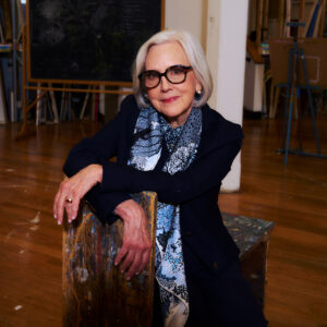 A woman with gray hair and glasses sits on a stool in an art studio. She is wearing a dark outfit with a patterned scarf. The background features a wooden floor and a chalkboard, along with art supplies and easels. She rests her arms casually on the stool, exuding a warm and approachable demeanor.