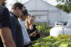 A group of four people stands in a garden setting, examining a young plant held by one individual. The people appear engaged in conversation about the plant, with expressions of curiosity and contemplation. Behind them, a greenhouse is visible, and various plants are seen growing in the foreground. A propane tank can be seen to the right. The scene suggests a learning or farming activity.