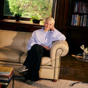 A smiling person with short light hair sits on a light beige sofa, resting their chin on one hand. They wear a loose blue shirt and black pants, paired with patterned shoes. The background features a window with greenery visible outside and a wooden bookshelf filled with books. A small glass table holds floral arrangements and a stack of colorful books. Soft lighting enhances the warm atmosphere of the setting.