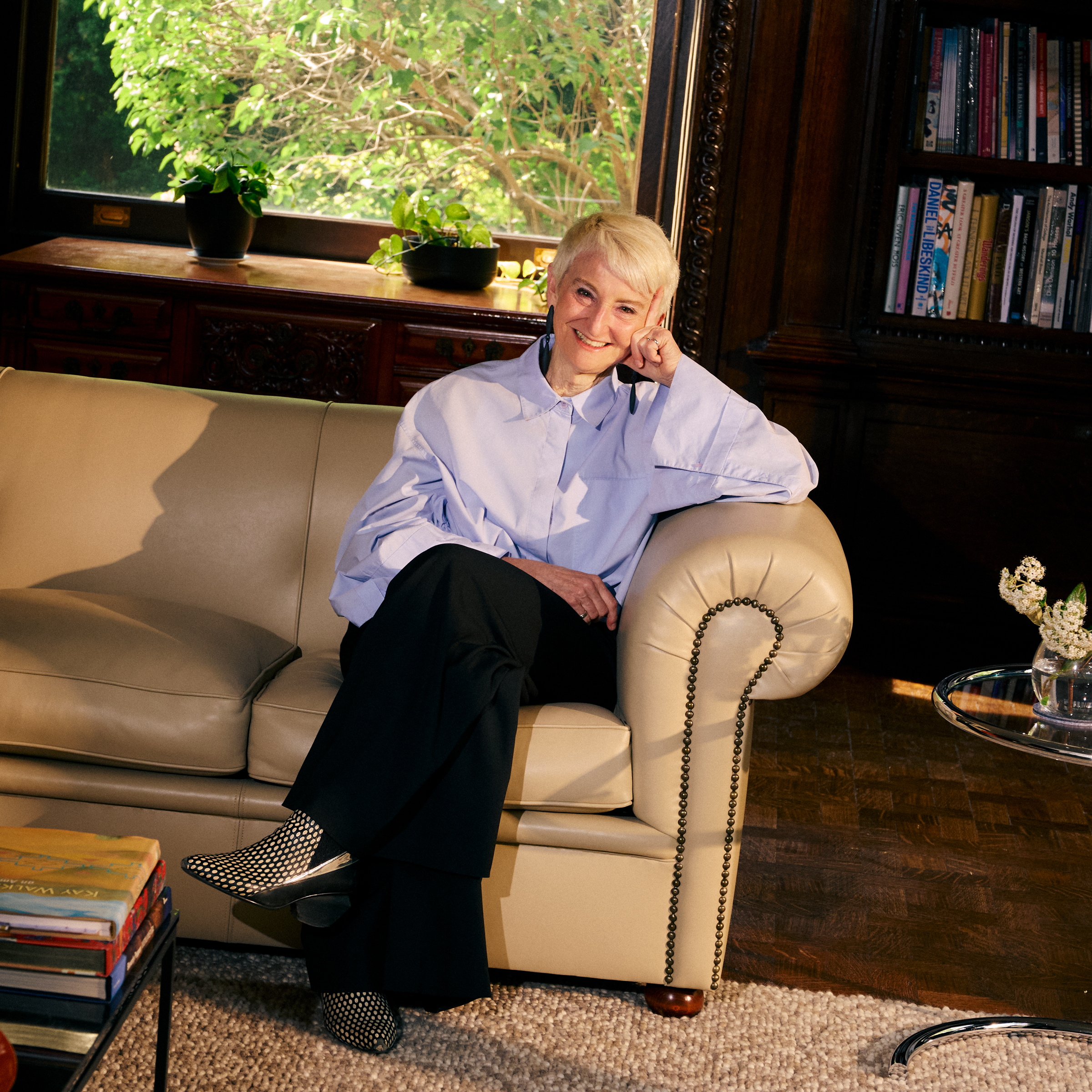 A smiling person with short light hair sits on a light beige sofa, resting their chin on one hand. They wear a loose blue shirt and black pants, paired with patterned shoes. The background features a window with greenery visible outside and a wooden bookshelf filled with books. A small glass table holds floral arrangements and a stack of colorful books. Soft lighting enhances the warm atmosphere of the setting.
