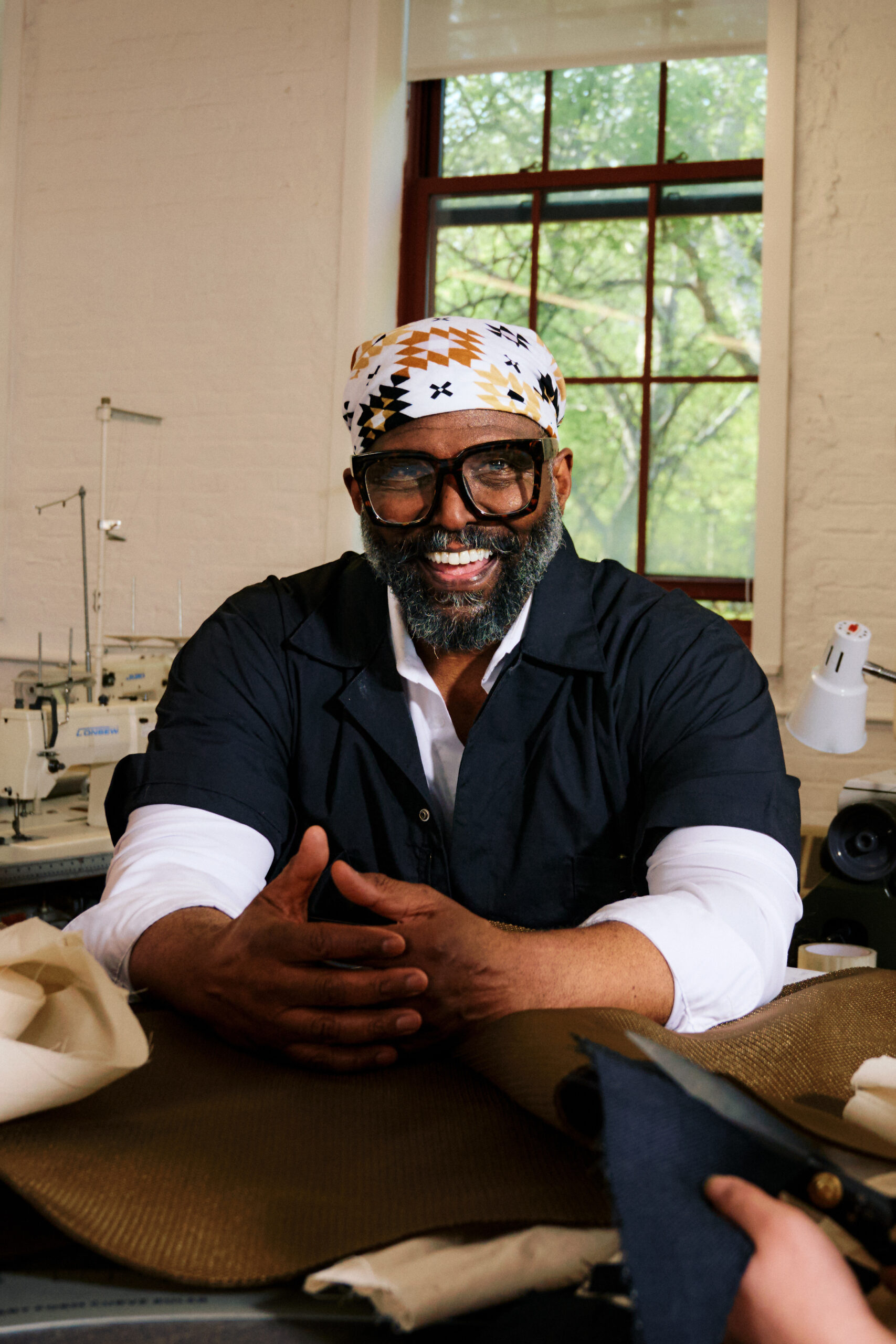 A smiling man with a beard and glasses sits at a work table, wearing a patterned headscarf and a dark shirt. He has his hands clasped in front of him, and there are various fabrics and sewing tools on the table. A sewing machine is visible in the background, along with a window showing green foliage outside.