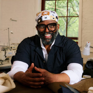A smiling man with a beard and glasses sits at a table, wearing a black shirt and a patterned bandana. His hands are placed in front of him, and he appears cheerful. Behind him are sewing machines and a window with greenery visible outside.