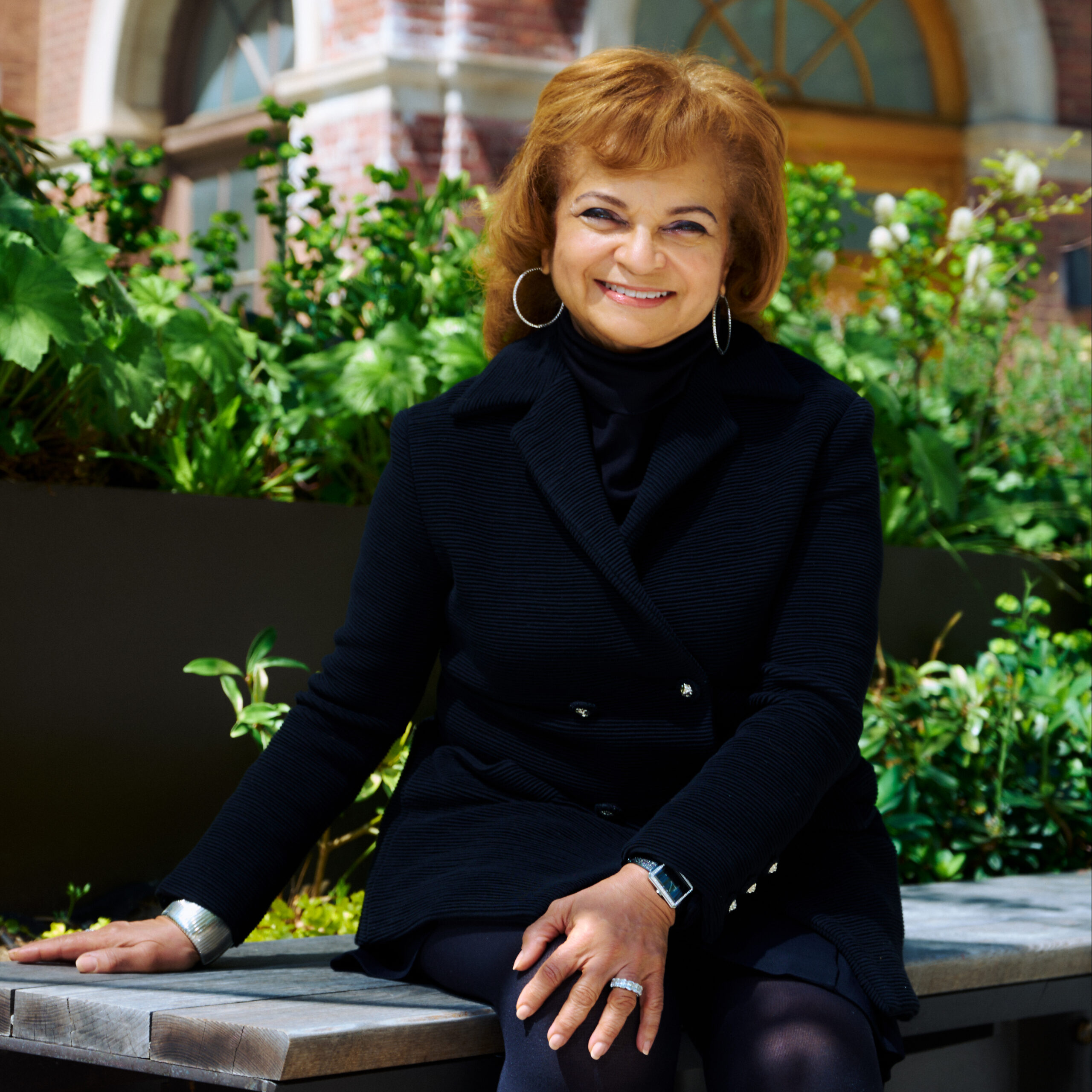 A woman with wavy brown hair, wearing a black coat and turtleneck, sits on a wooden bench surrounded by vibrant greenery. She smiles warmly at the camera, showcasing large earrings and a watch on her wrist. The background features a brick building with large windows, providing a bright and inviting atmosphere.