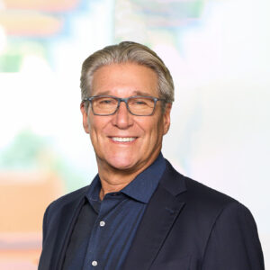 A man with gray hair and glasses is smiling at the camera. He is wearing a dark blazer over a collared shirt. The background is softly blurred with light colors, giving a professional appearance.