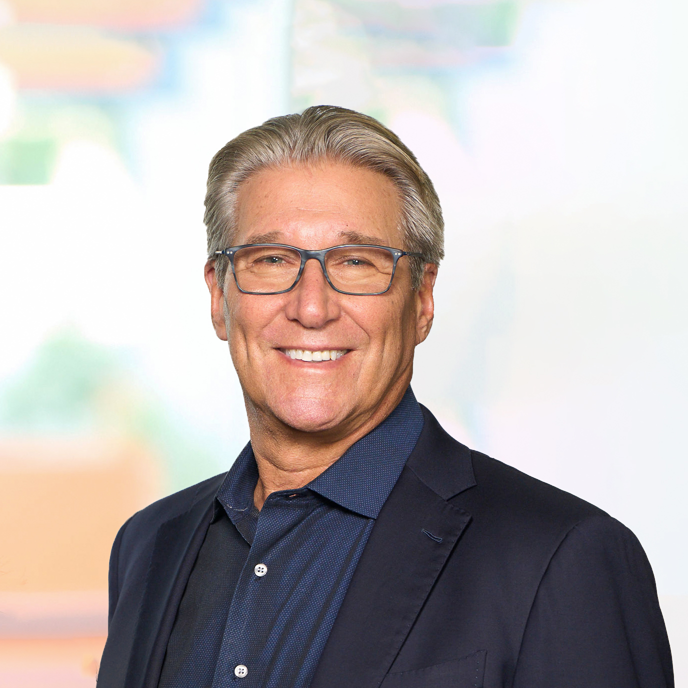 A man with gray hair and glasses is smiling at the camera. He is wearing a dark blazer over a collared shirt. The background is softly blurred with light colors, giving a professional appearance.
