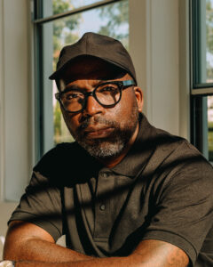 A man with a beard and glasses sits with his arms crossed, wearing a black cap and a dark polo shirt. Sunlight casts shadows across his face, highlighting his serious expression. The background features large windows with greenery visible outside.