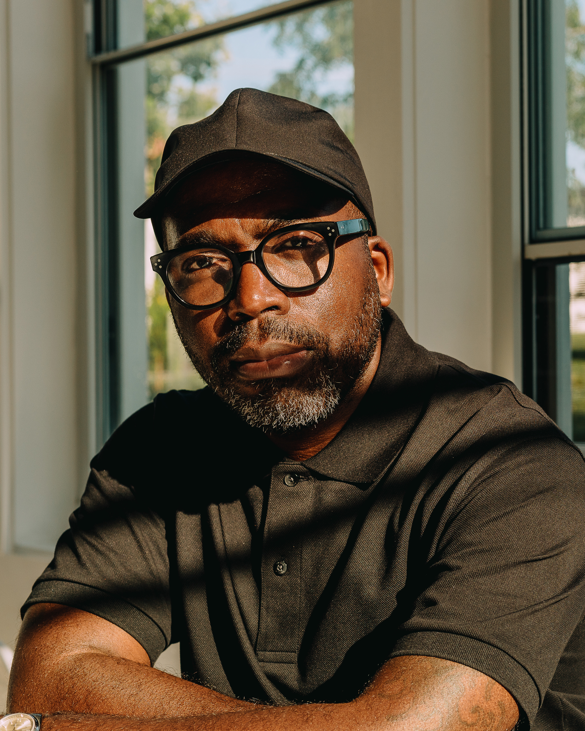 A man with a beard and glasses sits with his arms crossed, wearing a black cap and a dark polo shirt. Sunlight casts shadows across his face, highlighting his serious expression. The background features large windows with greenery visible outside.
