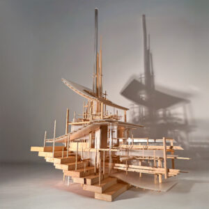 A detailed architectural model constructed from wood and transparent materials, featuring multiple levels and intricate staircases. It includes vertical elements and overhanging structures that create a dynamic silhouette, casting a shadow on the background. The overall design combines geometric forms with organic shapes, suggesting a contemporary architectural style. The lighting enhances the model's textures and depth.