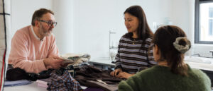 Three individuals are engaged in a discussion at a table covered with fabric and clothing. One person, wearing a pink sweatshirt and glasses, is holding a patterned fabric swatch while speaking. The second individual, in a striped sweater, is listening attentively with a slight smile. The third person, wearing a green sweater, is partially visible with her back to the camera, focused on the conversation. The setting appears to be a creative workspace.