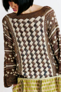 A person with a bob haircut is shown wearing a textured, oversized sweater with a geometric pattern in shades of brown and cream. The sweater has a wide neckline and long sleeves. Below the sweater, a green skirt with horizontal stripes and circular patterns is partially visible, adding a contrasting color to the outfit. The background is softly blurred, focusing on the clothing details.