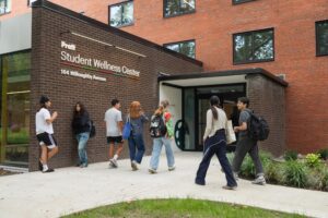 A group of diverse students walks towards the entrance of the Pratt Student Wellness Center, which features a modern design and large windows. The building is made of dark brick, with the center's name prominently displayed. Some students appear to be engaging in conversation while others walk towards the door. Green grass and small plants are visible in the foreground, adding to the inviting atmosphere.