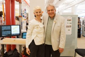 A woman and a man stand smiling closely together in a brightly lit exhibition space. The woman has short, platinum blonde hair and wears a white jacket, while the man has gray hair and is dressed in a light-colored blazer over a green shirt. Both are wearing yellow buttons on their clothing. In the background, there are display boards and people engaged in conversation.