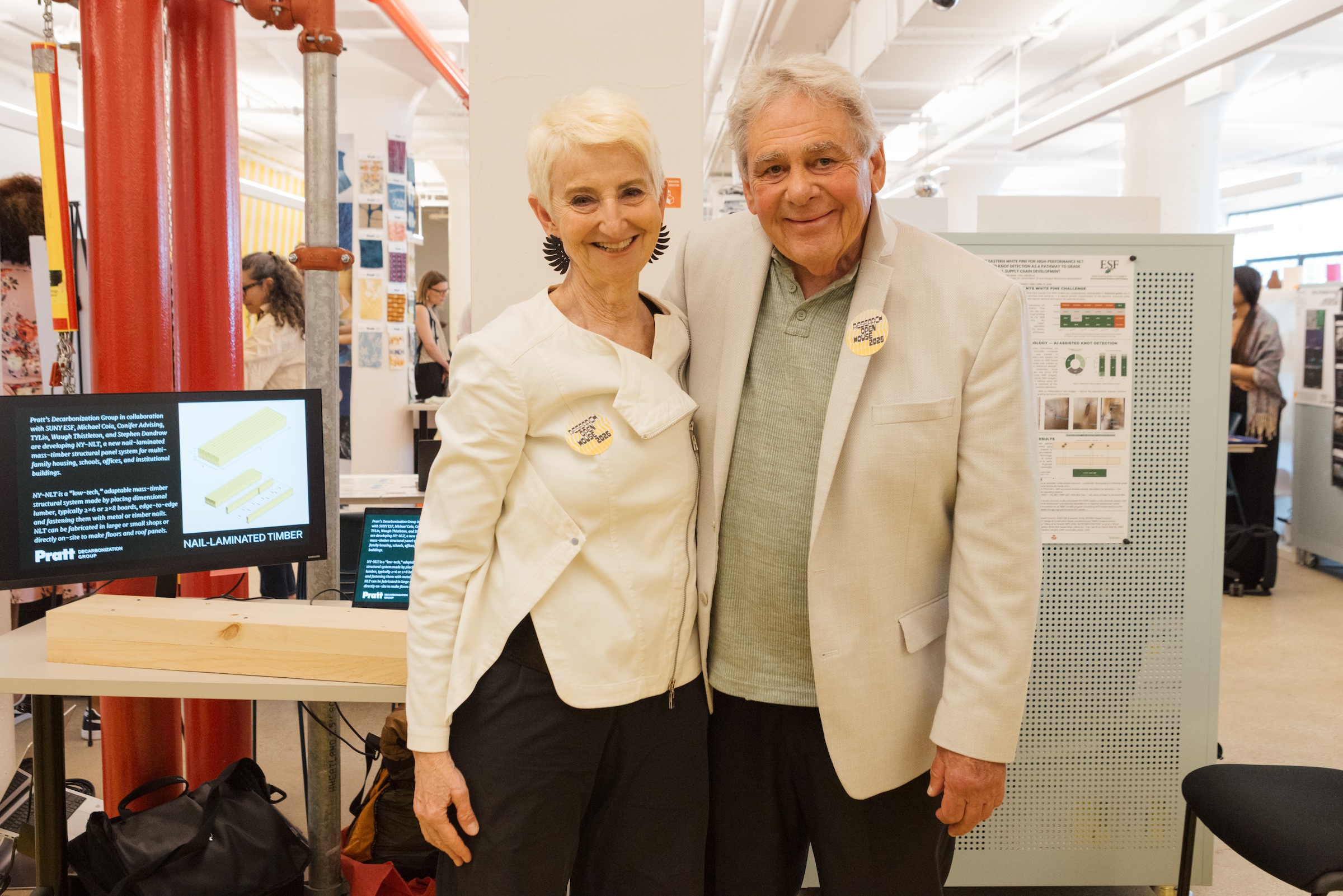 A woman and a man stand smiling closely together in a brightly lit exhibition space. The woman has short, platinum blonde hair and wears a white jacket, while the man has gray hair and is dressed in a light-colored blazer over a green shirt. Both are wearing yellow buttons on their clothing. In the background, there are display boards and people engaged in conversation.