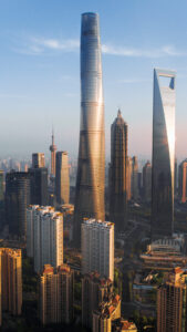 A skyline view of Shanghai featuring several skyscrapers including the Shanghai Tower, which is the tallest, with a spiraling design. Nearby are the Jin Mao Tower and the Shanghai World Financial Center, distinguished by their unique shapes. The foreground shows modern residential buildings, and the background displays a clear blue sky.