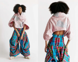 A model poses wearing an oversized, pink, velvet top with embroidered details on the back. The top has a loose, draped silhouette. The model’s lower half is adorned with baggy, vibrant plaid trousers in blue, red, and green colors, featuring large pockets. Soft, fluffy slippers complete the outfit. The background is a plain white, highlighting the fashion ensemble.