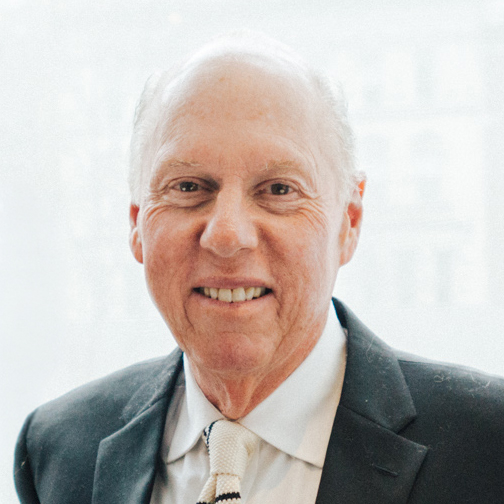 A smiling older man with gray hair is wearing a dark suit and a white shirt with a tie. He is standing in front of a light, blurred background, suggesting an indoor setting with natural light.