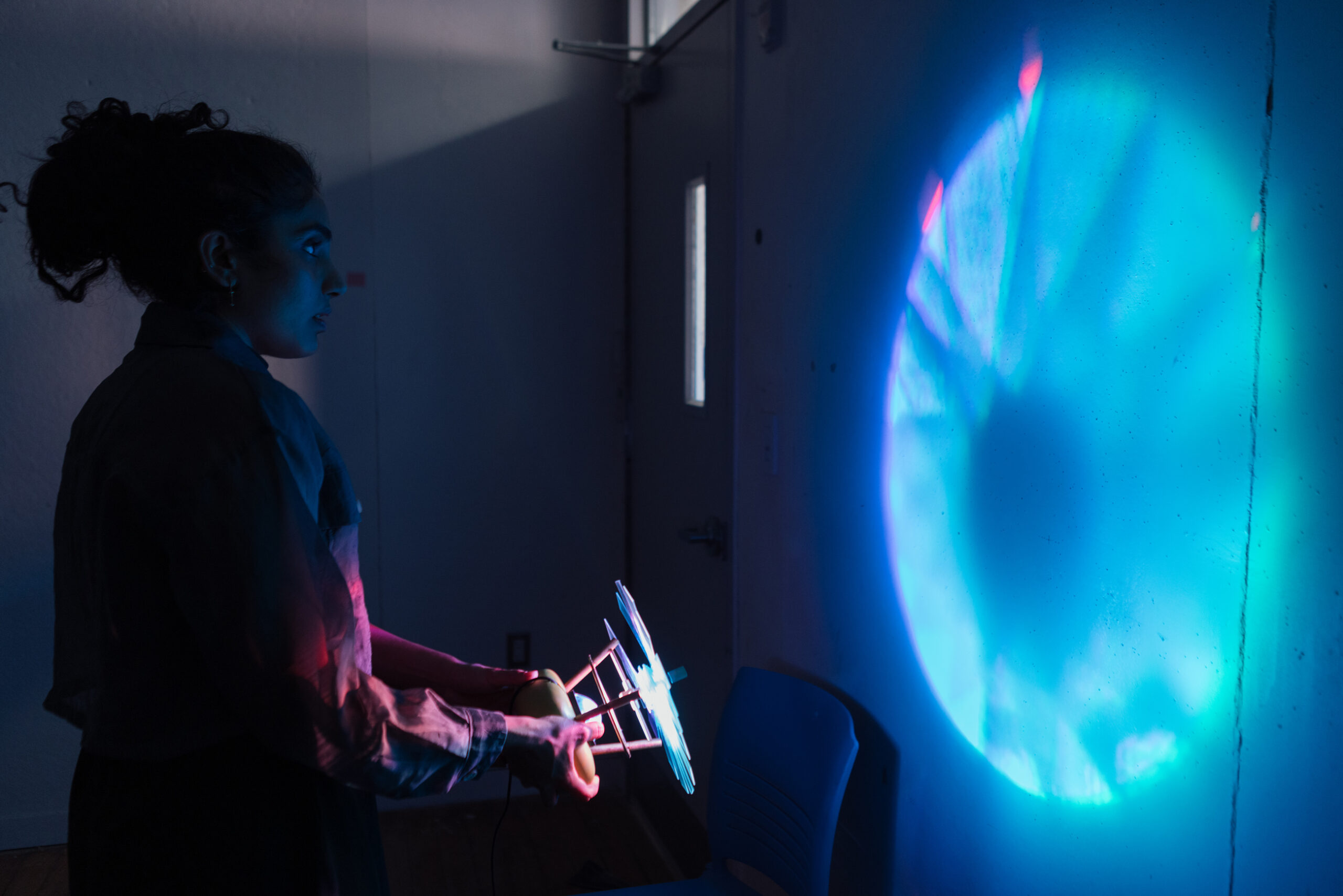 A person with curly hair stands in a dimly lit room, holding a device that projects colorful light patterns onto a wall. The projection displays intricate, swirling blue and turquoise shapes, while the person's profile is illuminated by the glow. There is a chair in the foreground against a plain wall, and a door with a window is visible in the background.