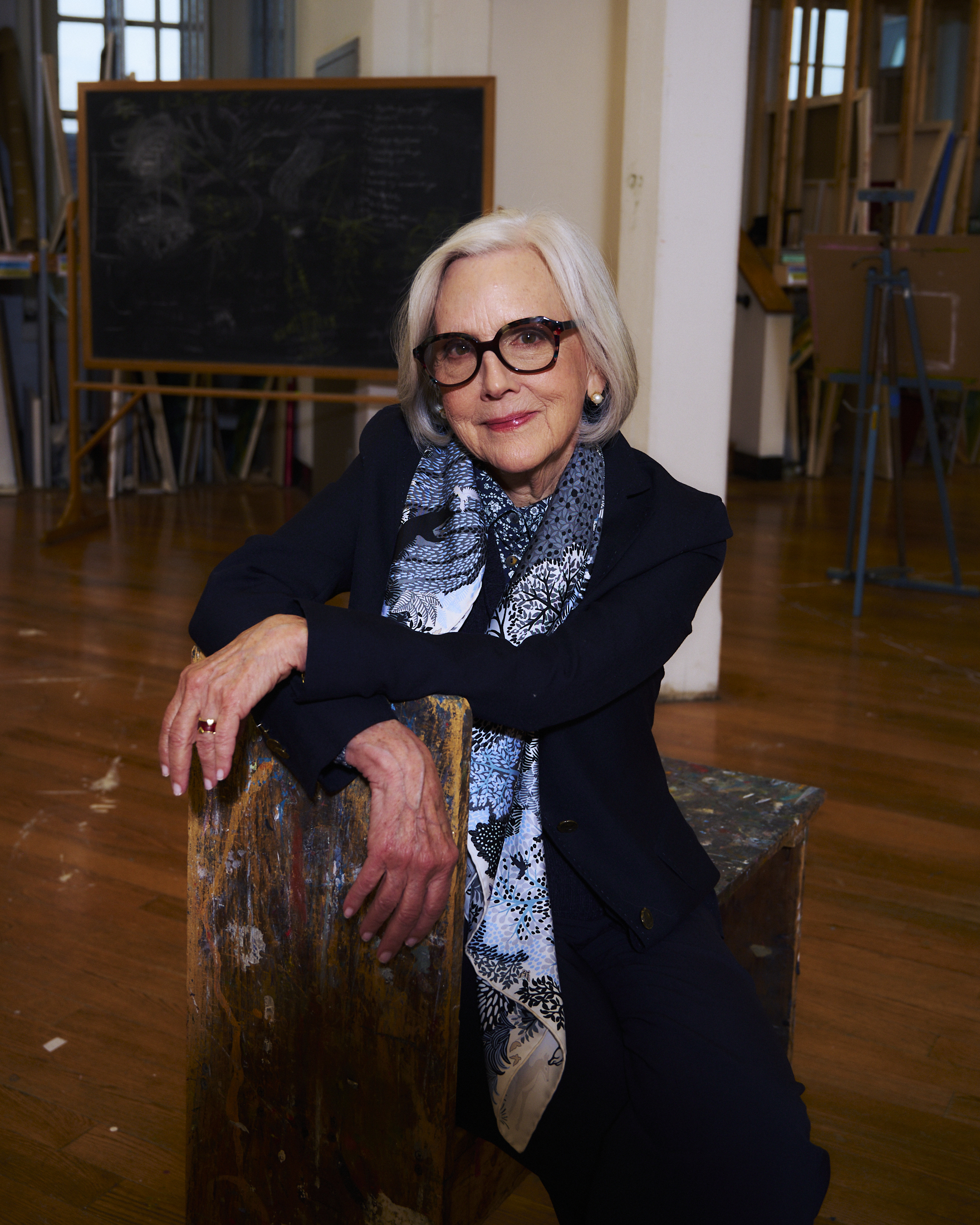 A woman with silver hair and glasses smiles while seated on a rustic wooden stool. She wears a dark blazer and a patterned scarf. Behind her is a chalkboard with writing and drawings, and the room features art supplies and easels, indicating a creative workspace. The wooden floor reflects warm tones.