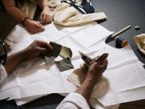 A close-up of two hands working with fabric swatches and pattern pieces on a table. One hand holds several cut pieces of fabric, including gray and green materials, while the other hand is near a set of fabric patterns and sewing tools, including scissors and a spool of thread. The background shows light-colored fabric and sewing notations.