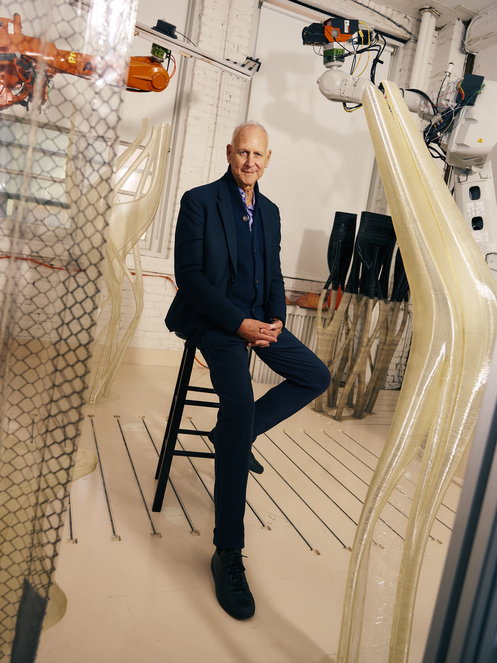 A seated man in a dark suit and black shoes poses confidently in a studio filled with robotic arms and translucent sculptural designs. The environment has a modern, industrial feel, with intricate structures made of flexible materials in the background, surrounded by a white brick wall.