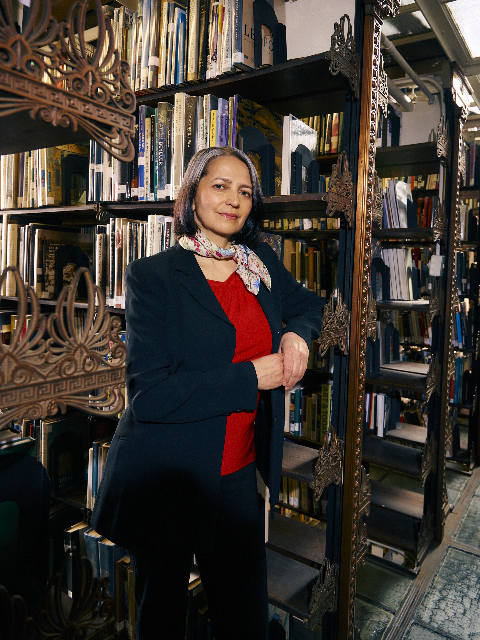 A woman with long, dark hair stands confidently in a library surrounded by bookshelves filled with various books. She wears a red top and a black blazer, with a floral scarf around her neck. Her arms are crossed as she leans against a shelf, looking directly at the camera with a slight smile. The setting is warmly lit, creating an inviting atmosphere.